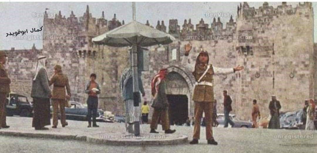 Traffic officer directing vehicles near a stone building with arched entrance and large walls. People on the street.