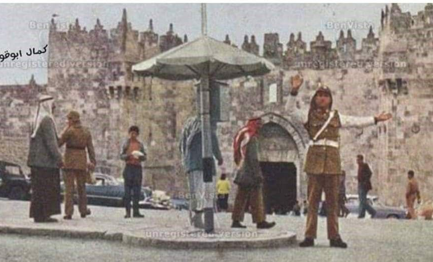 Traffic policeman directs vehicles near a stone gate in Jerusalem, people walking by.