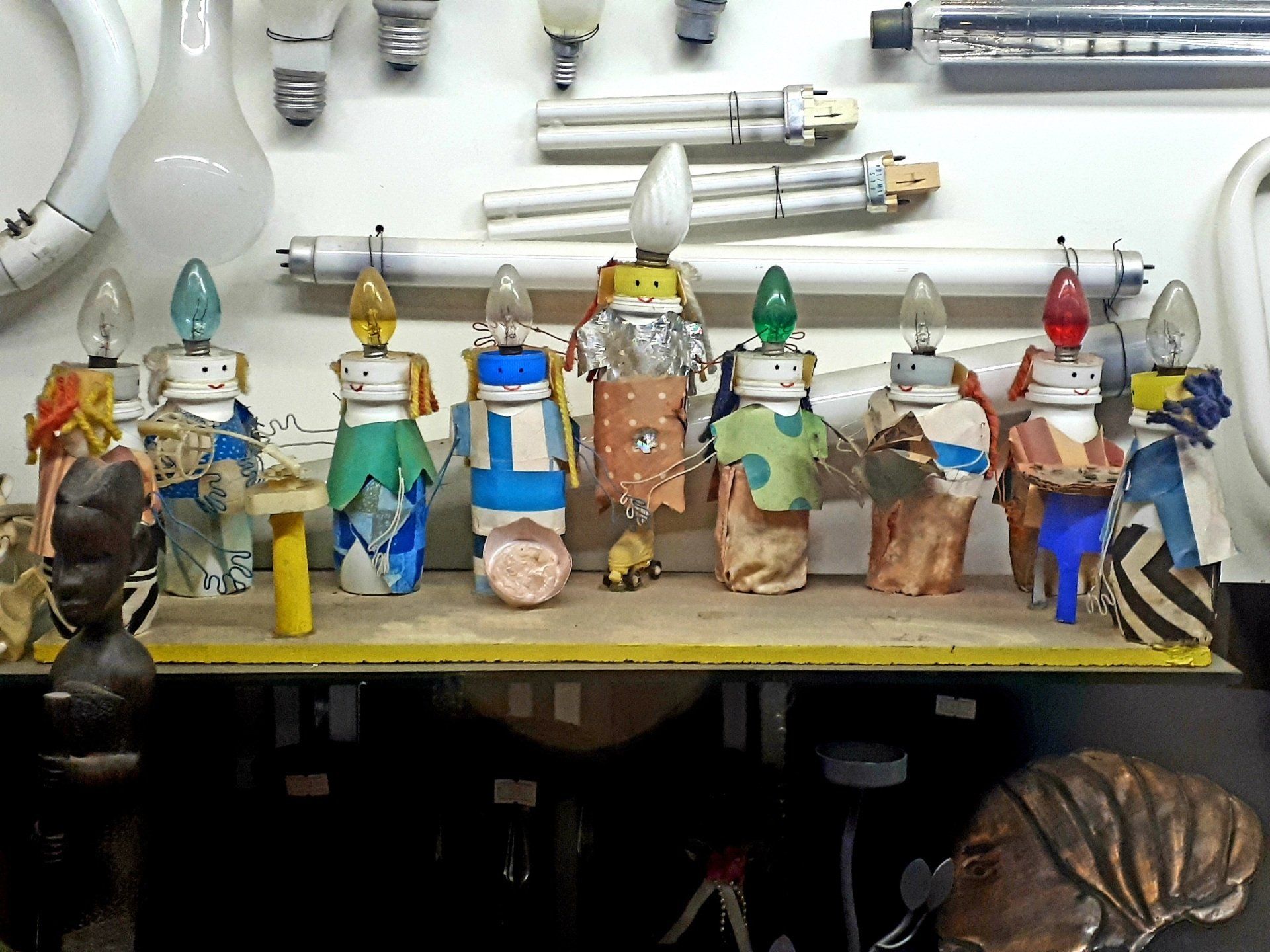 Collection of colorful art figures made from lightbulbs, atop a shelf with various overhead light fixtures in background.
