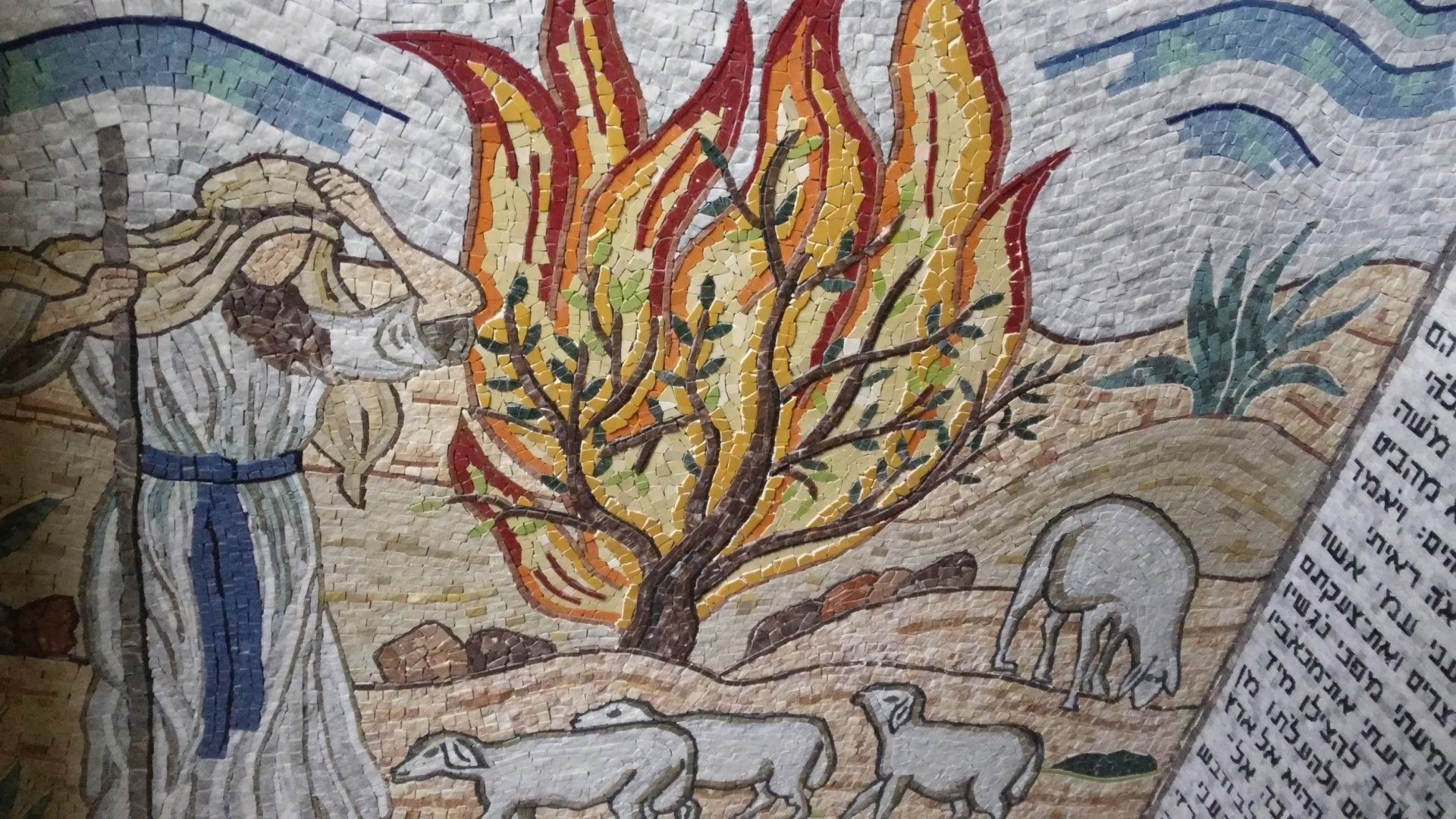 Mosaic of Moses and the burning bush. Moses in white robes, bush ablaze, sheep grazing, and text on the side.