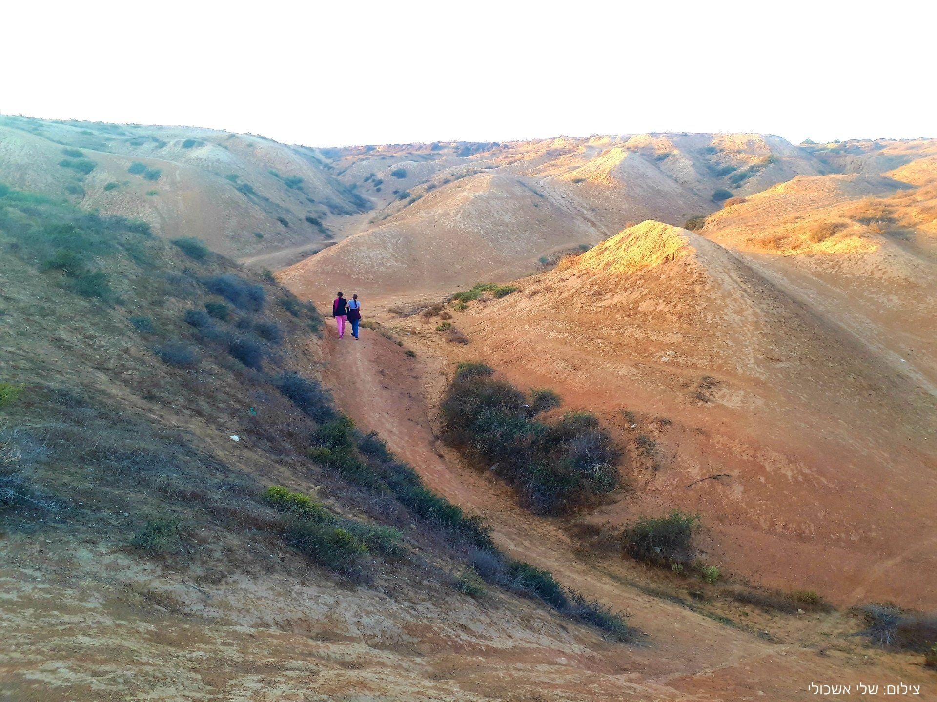 Two people hike a dirt trail through brown, hilly landscape.