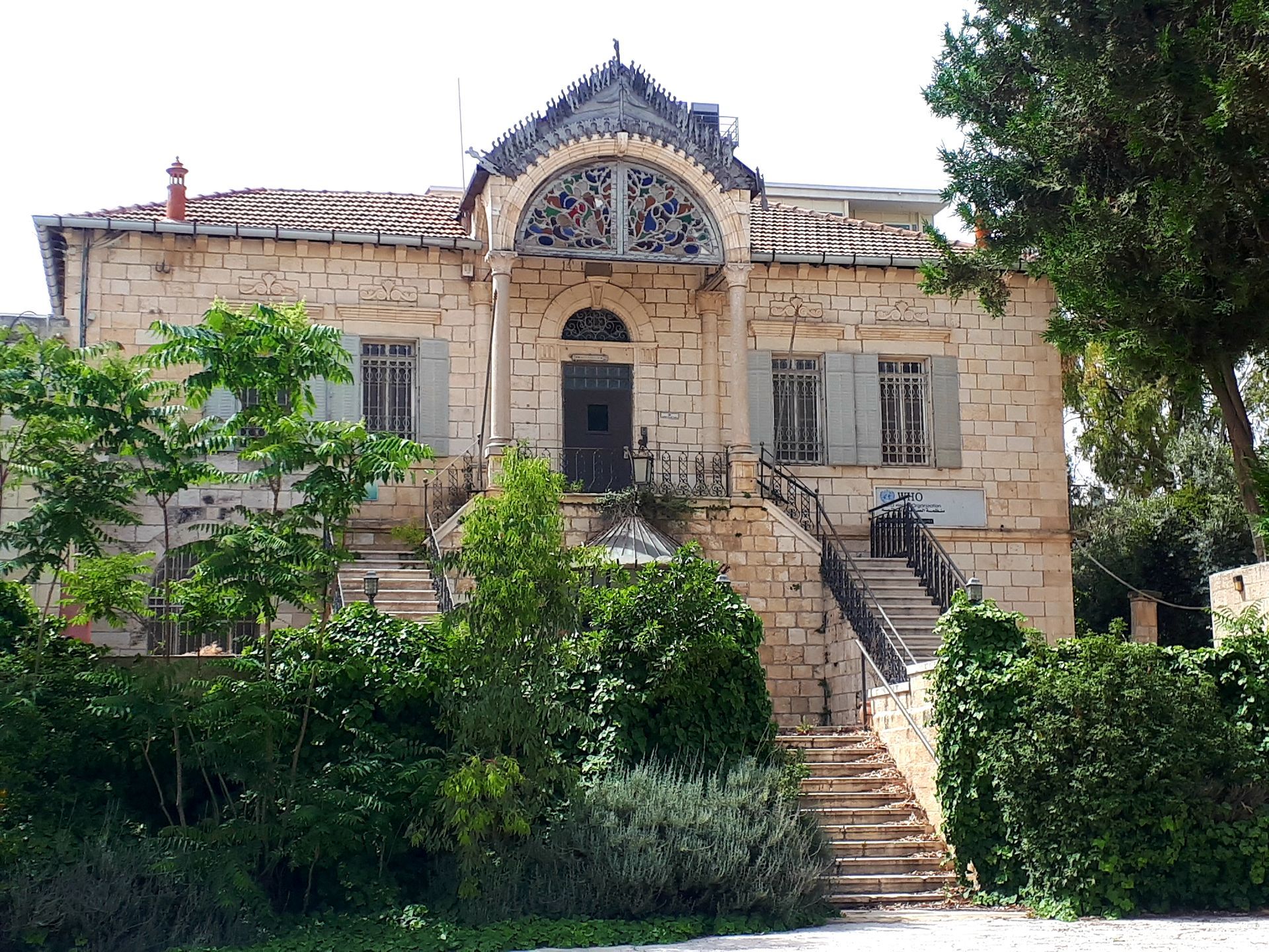 Stone house with arched entrance and exterior staircase, surrounded by greenery.