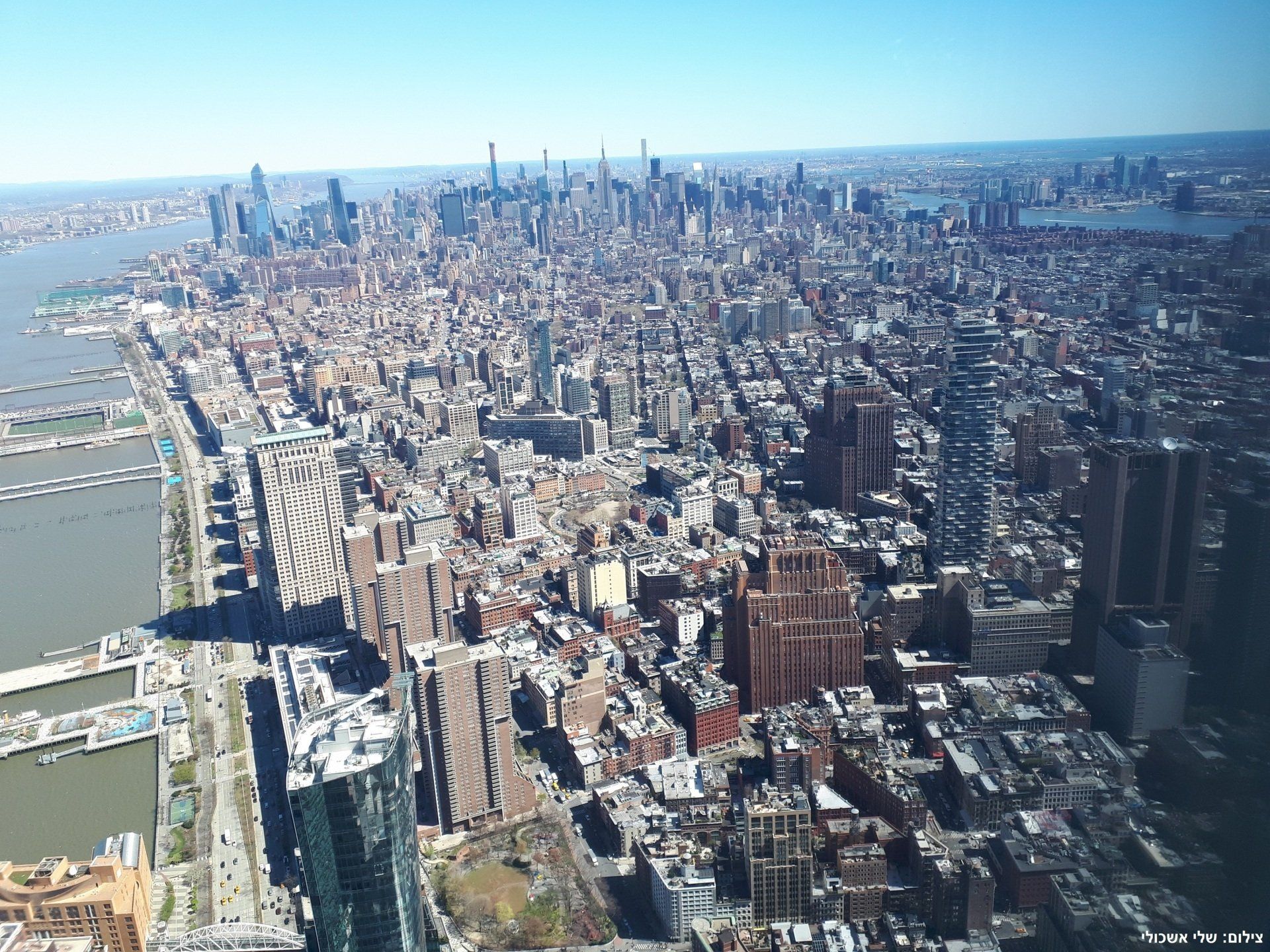 Aerial view of a densely packed city skyline with numerous skyscrapers, surrounded by water under a clear blue sky.