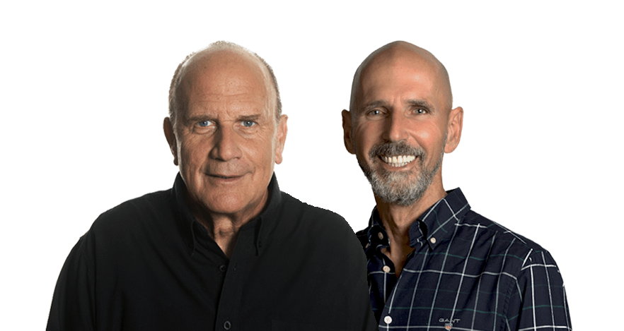 Two bald men smiling, posing closely together against a white backdrop. One in black, the other in a plaid shirt.