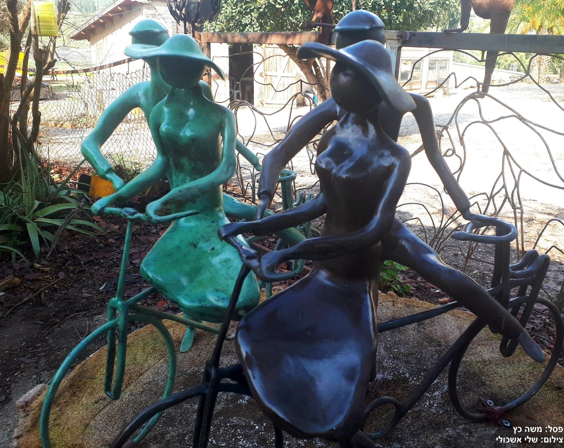 Bronze sculptures of figures on bicycles; green and dark bronze. Park setting, sunny day.