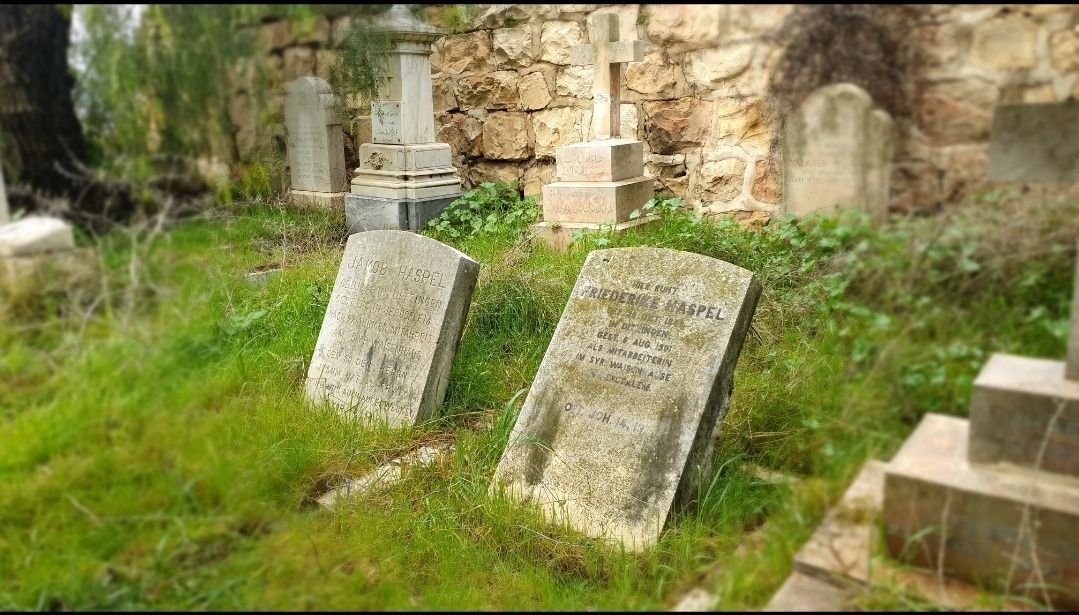 Weathered tombstones in a grassy cemetery, some leaning, with a stone wall background.