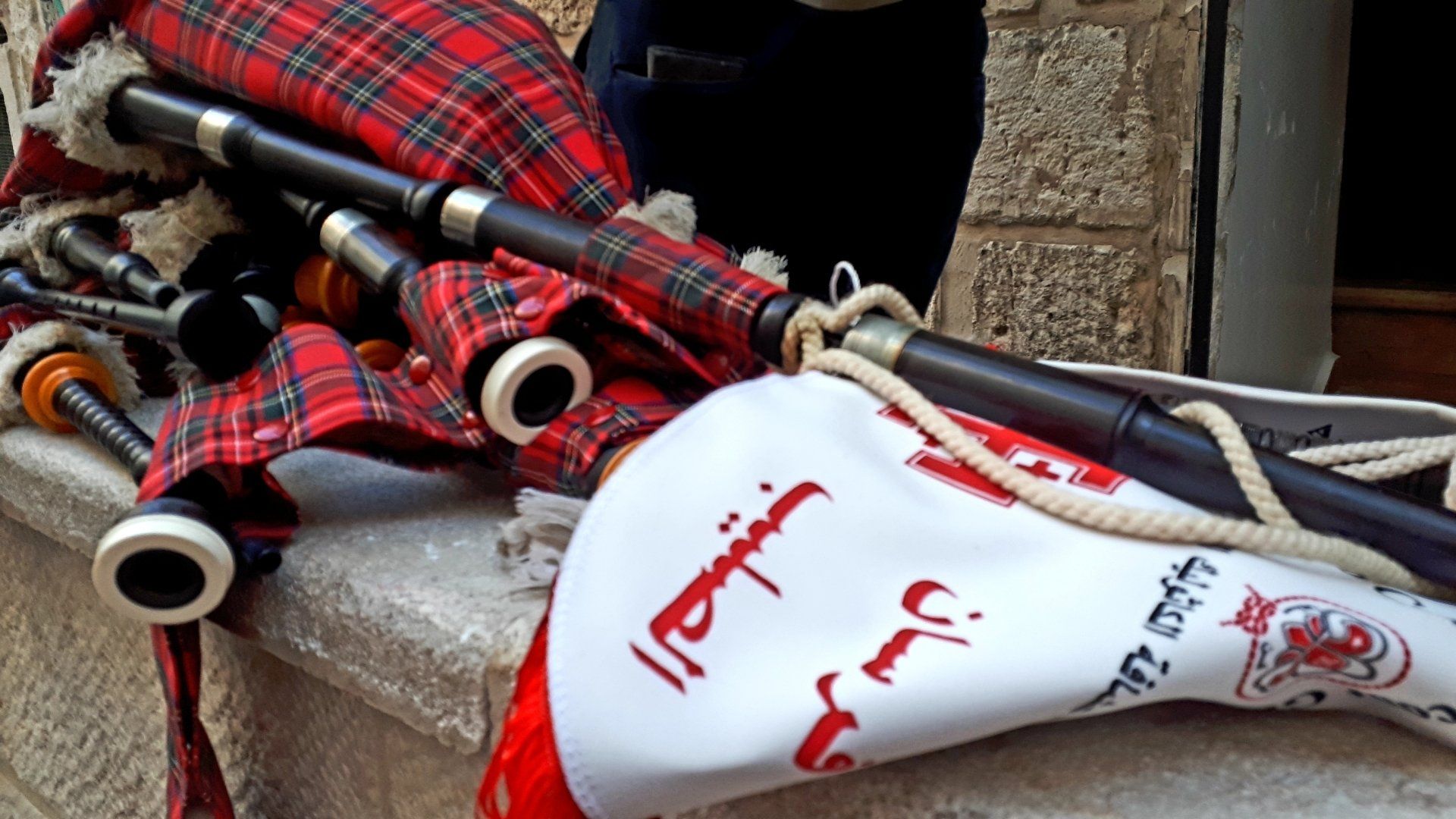 Bagpipes in red plaid tartan with a white bag featuring Arabic text.