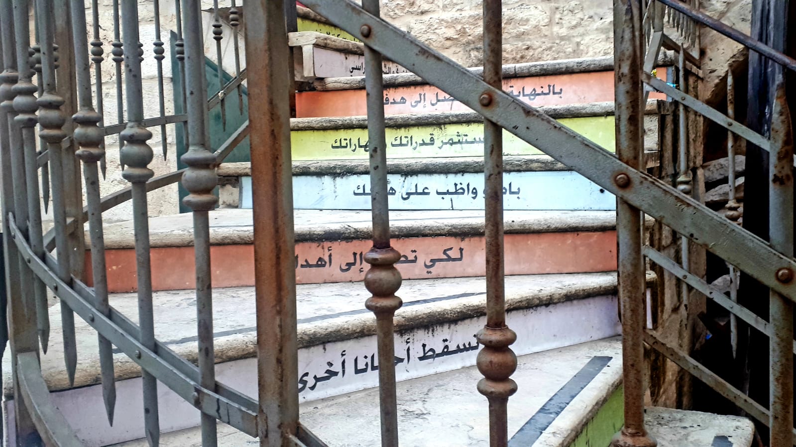 Steps with colorful risers and Arabic script, viewed through a wrought iron fence.