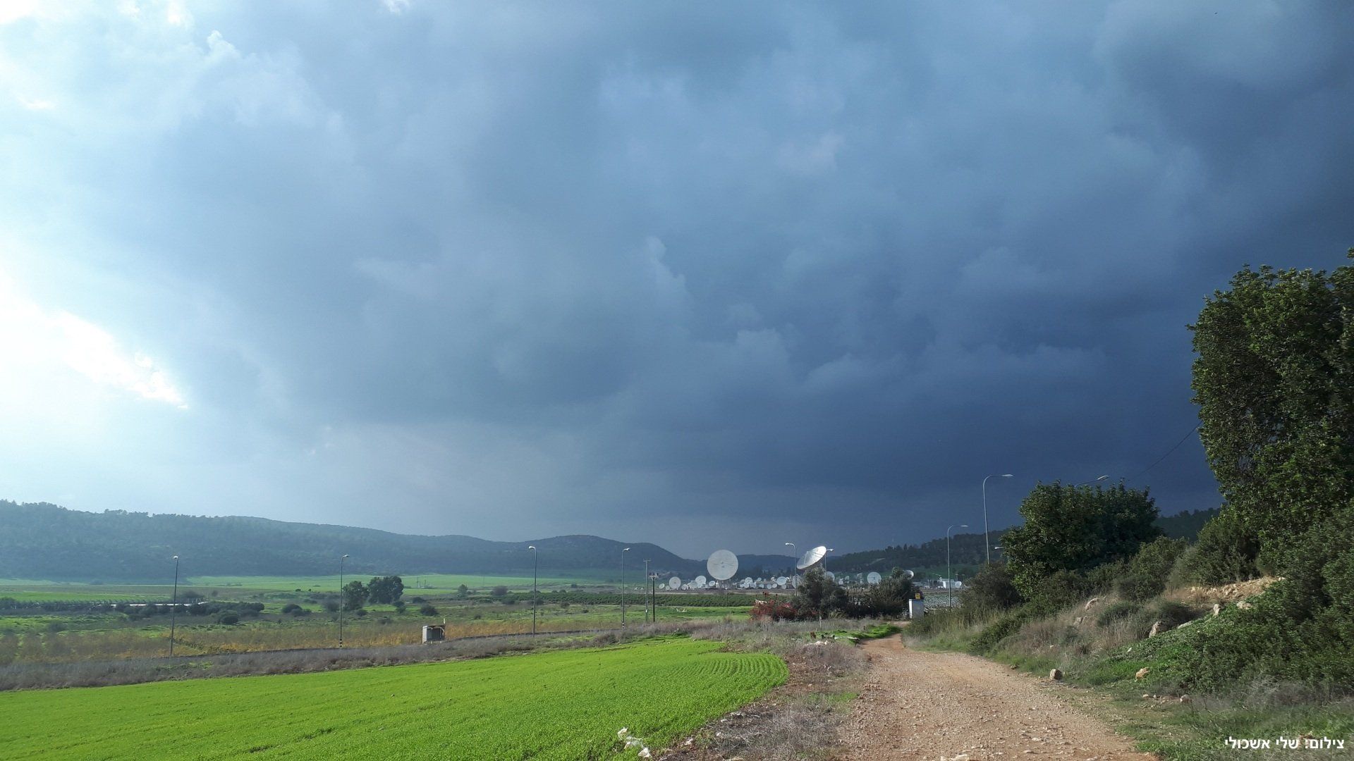 Dark storm clouds over a rural landscape with fields, dirt road, trees, and buildings.