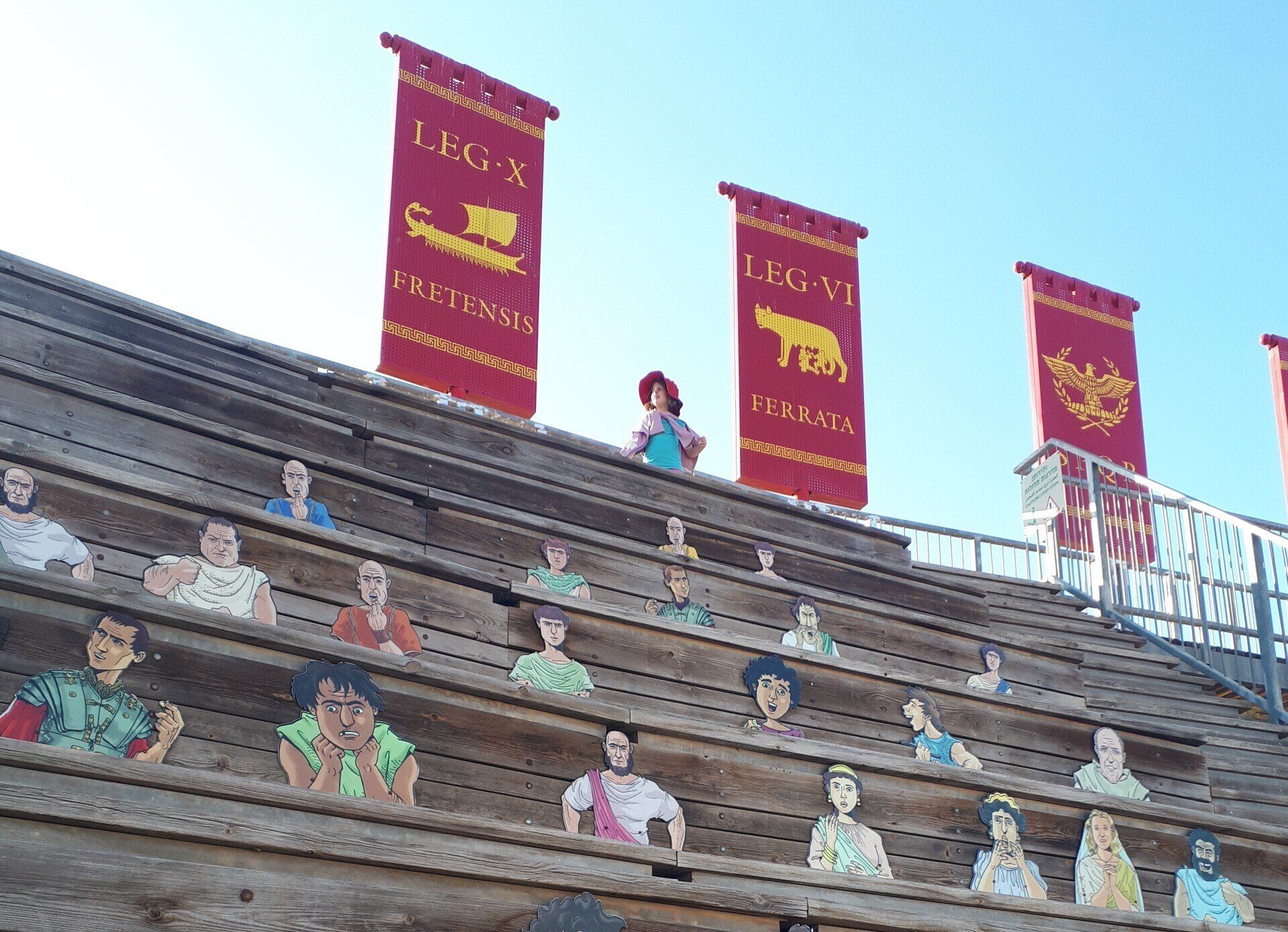 Wooden bleachers with cutouts of people, banners with Roman legion symbols, and a person standing on the top row.