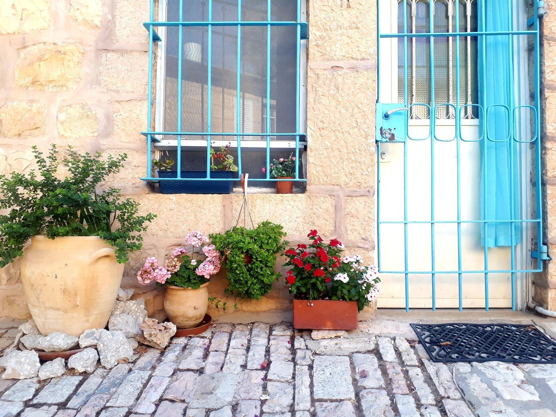 Stone building exterior with blue barred windows and door, various potted flowers on a brick patio.