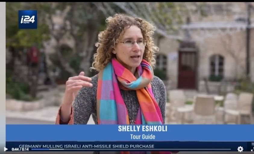 Shelly Eshkoli, a tour guide, speaks outdoors with a colorful scarf on. The i24 logo is in the corner.