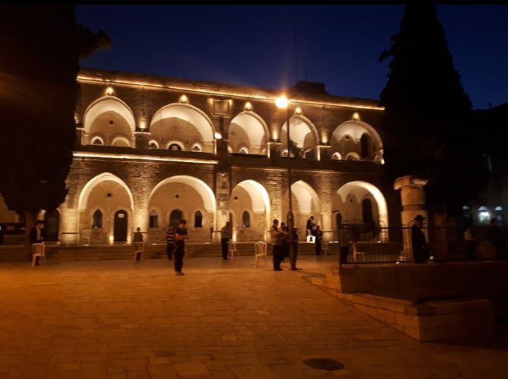 Night view of a stone building with arched windows, lit up. People are in the courtyard.