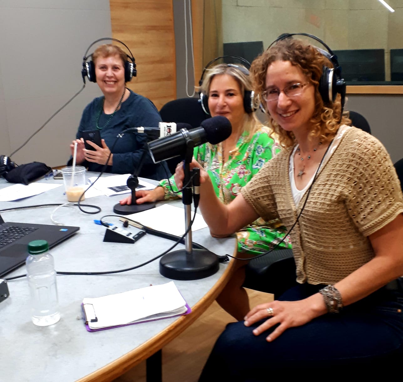 Three women in a recording studio, wearing headphones and speaking into microphones. Smiling at the camera.