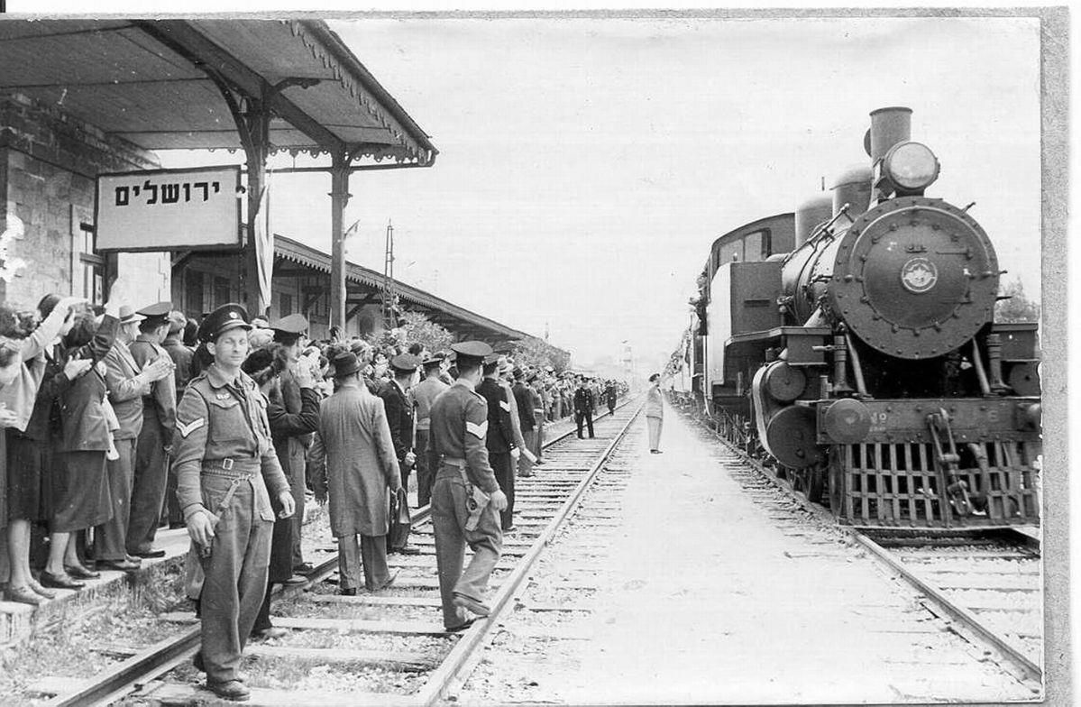 Steam train at a Jerusalem train station, people waving, soldiers on the tracks, sign in Hebrew.