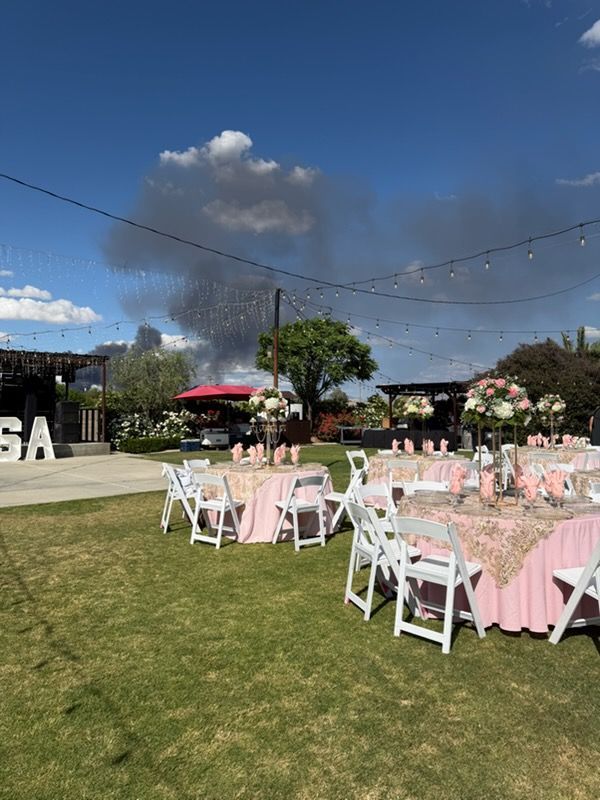 Outdoor lawn reception with pink tablecloths and white chairs under a blue sky, with smoke or fireworks in the distance
