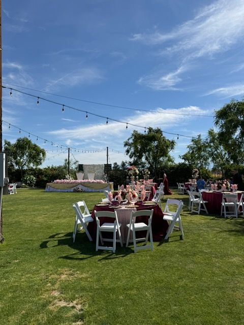 Outdoor reception with round tables, white chairs, and string lights on a grassy lawn under a blue sky.