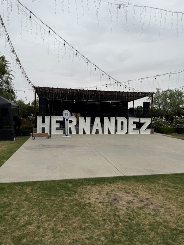 Large white “HERNANDEZ” letters on a concrete patio under string lights, with a covered stage behind.
