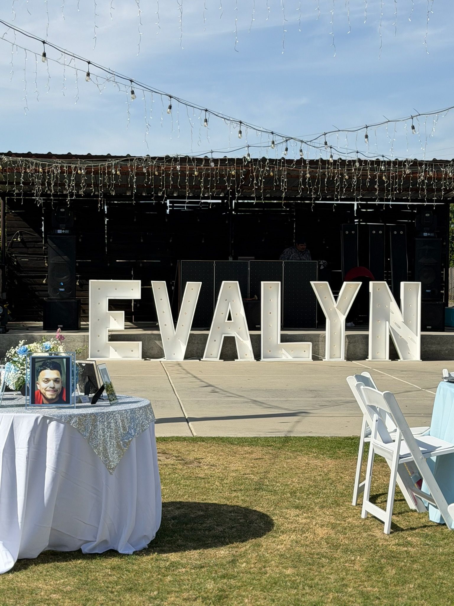 Outdoor event with large white “EVALYN” letters, decorated tables, and white chairs on a lawn under string lights