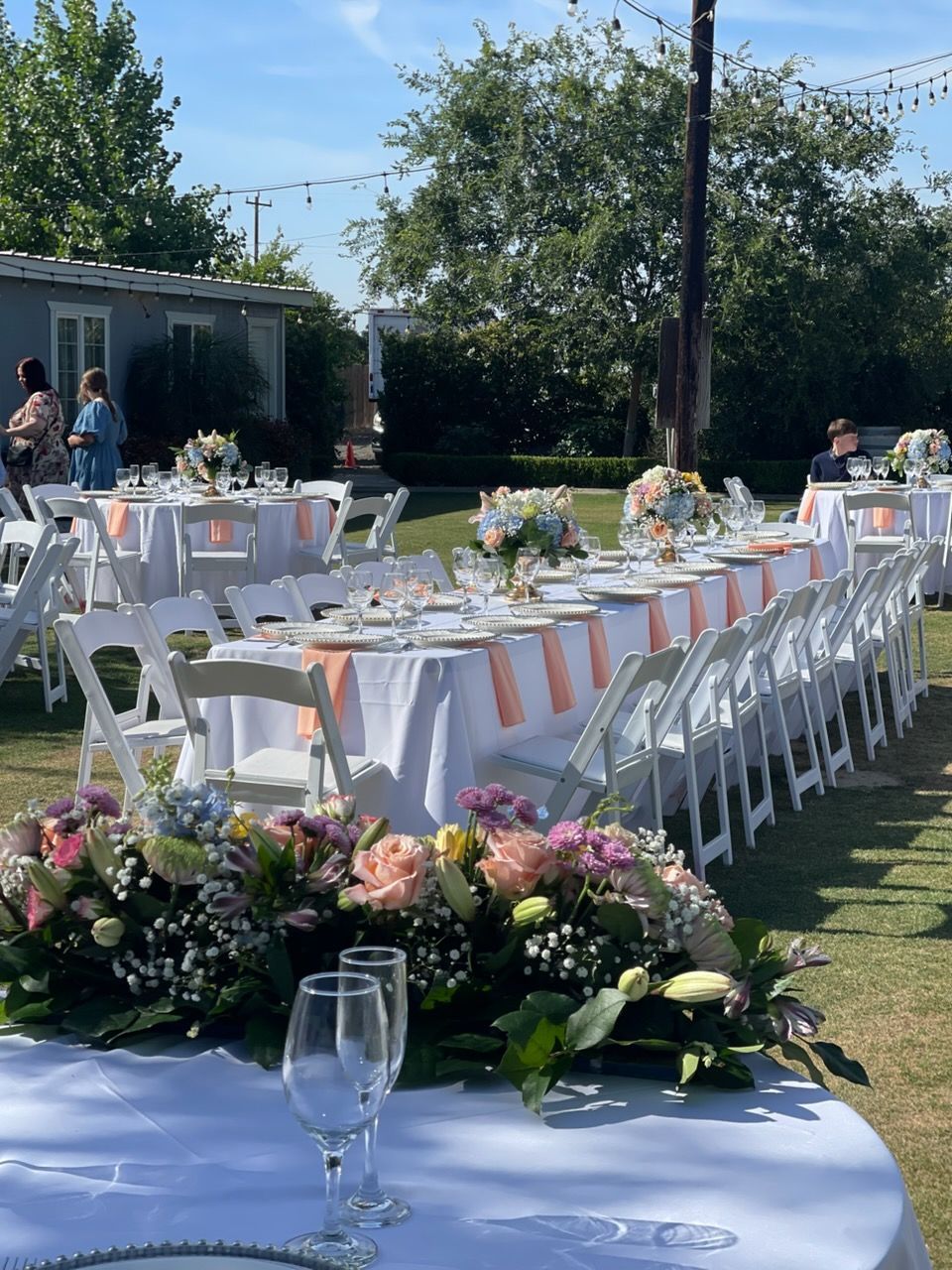 Outdoor wedding reception tables with white chairs, pink floral centerpieces, and guests in the background