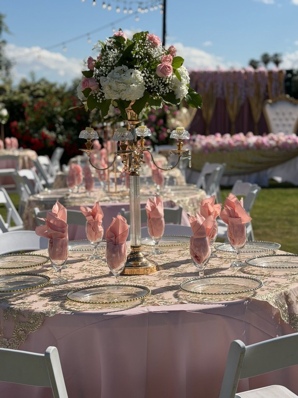 Round outdoor reception table with pink napkins and a floral centerpiece in a garden setting