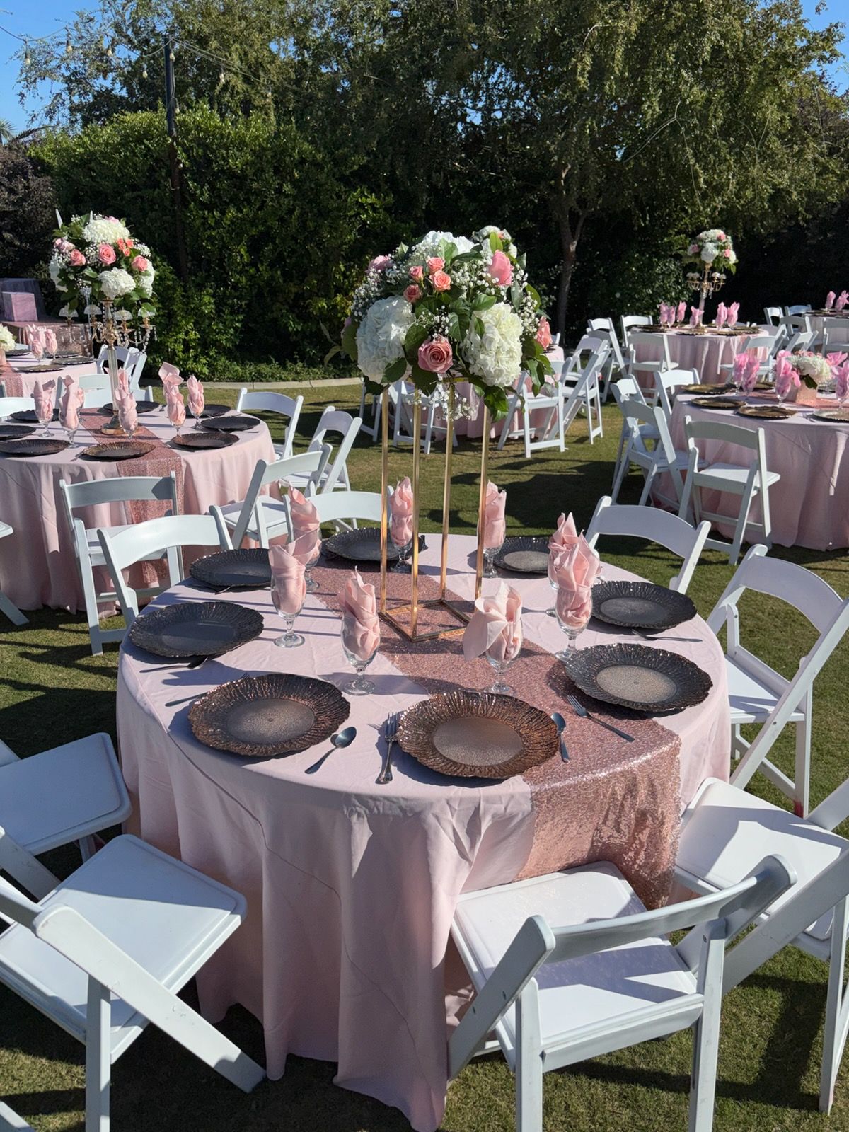 Round banquet tables set outdoors with pink tablecloths, white chairs, and floral centerpieces on a lawn.