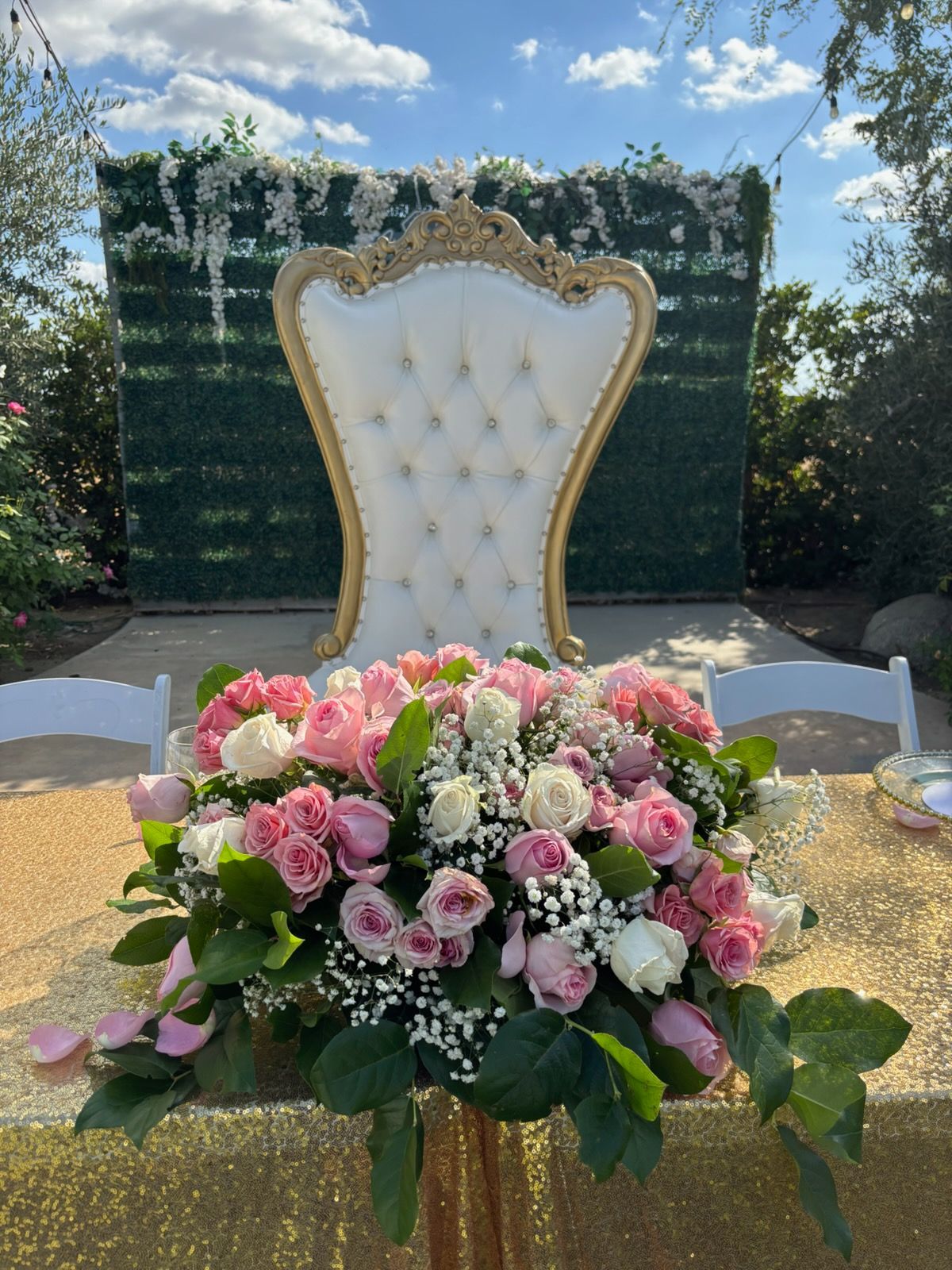 White tufted chair behind a large pink floral arrangement outdoors under a blue sky.