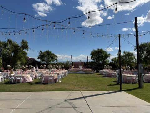 Outdoor wedding reception with white chairs, string lights, and tables on a grassy lawn under a blue sky