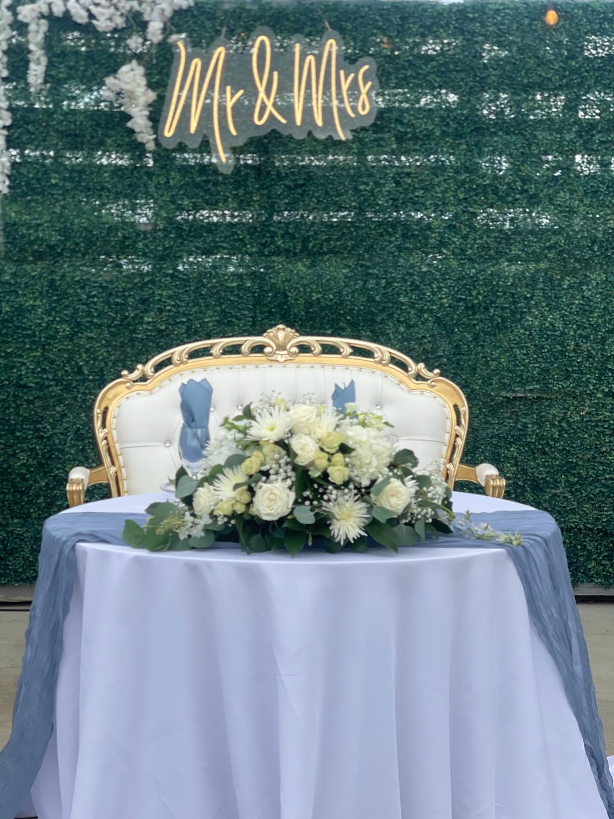 Decorated wedding sweetheart table with floral arrangement, gold chair, and “Mr & Mrs” sign on green backdrop