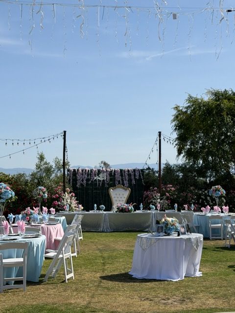 Outdoor wedding reception setup with white chairs, round tables, pink decor, and a floral stage on a lawn