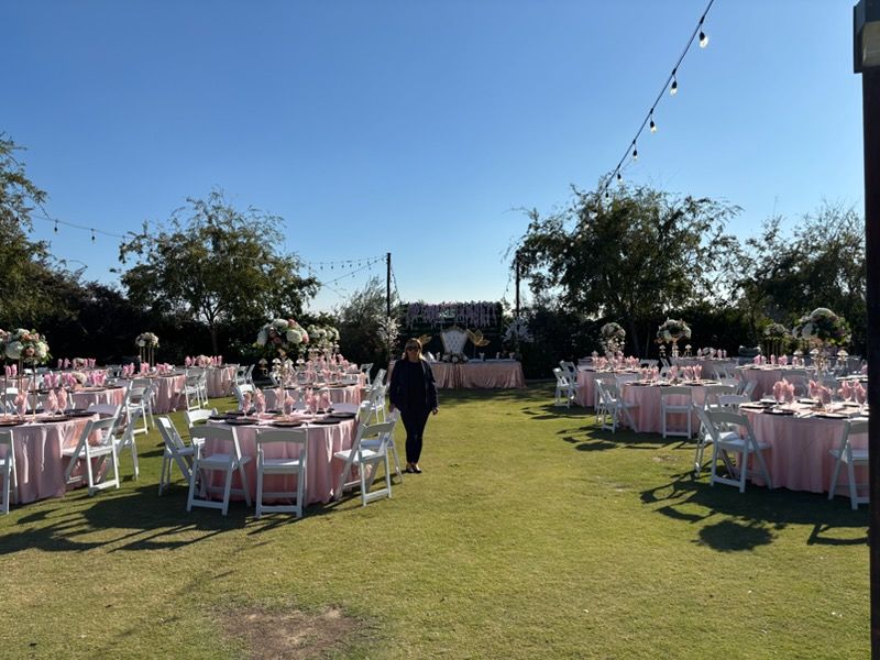 Outdoor event setup with pink chairs and tables on a grassy lawn, with string lights overhead and a stage in back