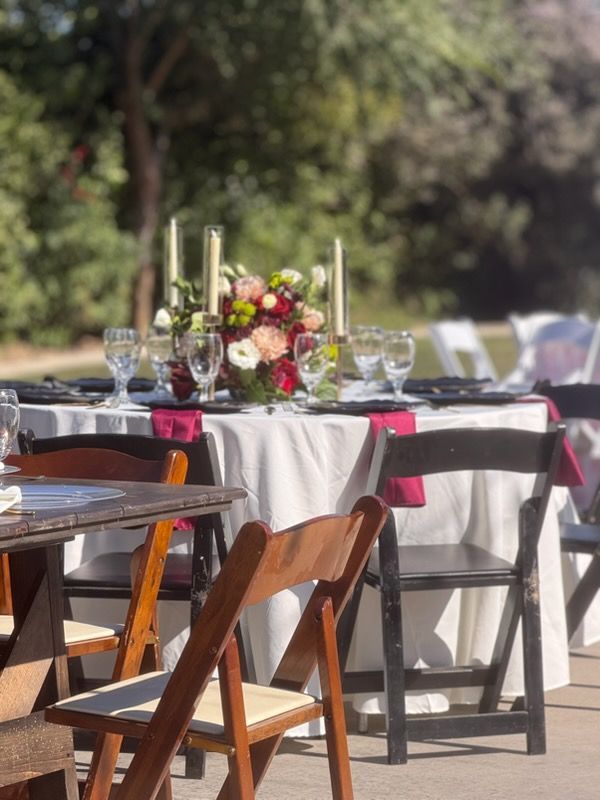 Outdoor event tables with white tablecloths, dark chairs, and floral centerpieces in a sunny garden setting