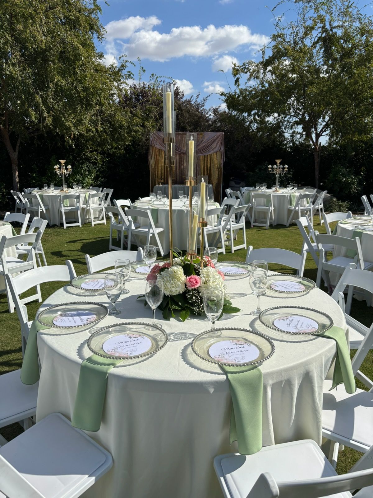 Outdoor banquet table set with white chairs, pale green linens, and floral centerpiece under trees