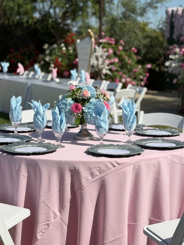 Round outdoor table set for a reception, with pink tablecloth, blue glassware, floral centerpiece, and white chairs