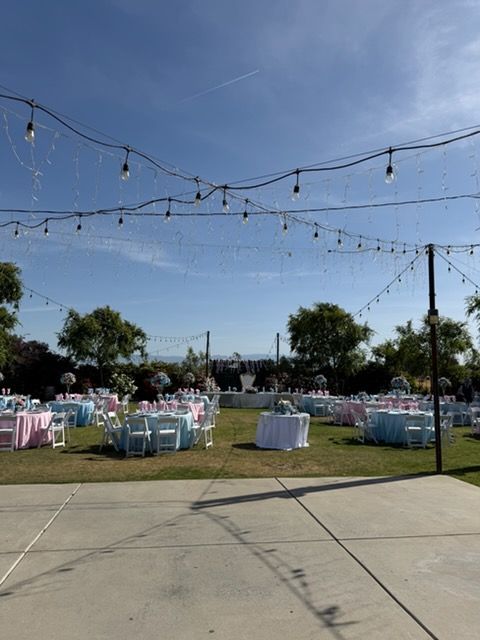 Outdoor reception setup with white-clothed tables, pastel chairs, and string lights on a grassy lawn