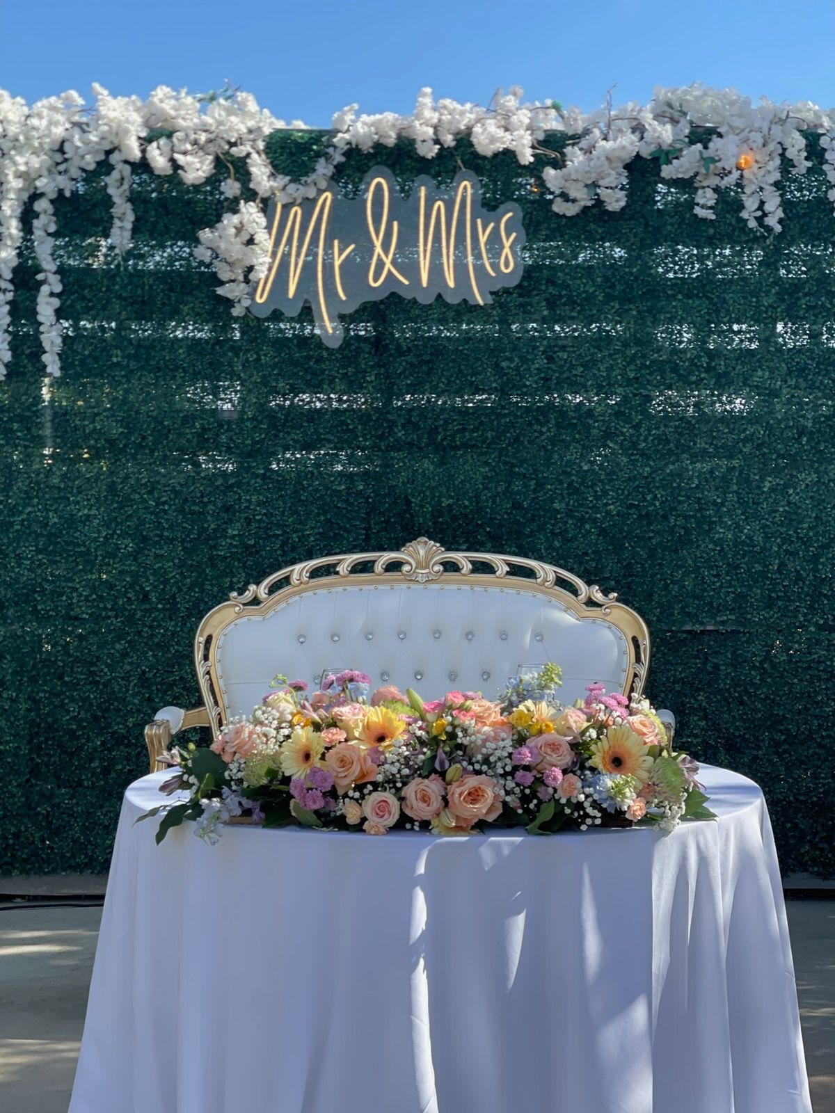 Floral wedding backdrop with gold Mr & Mrs sign, white loveseat, and flower-covered table