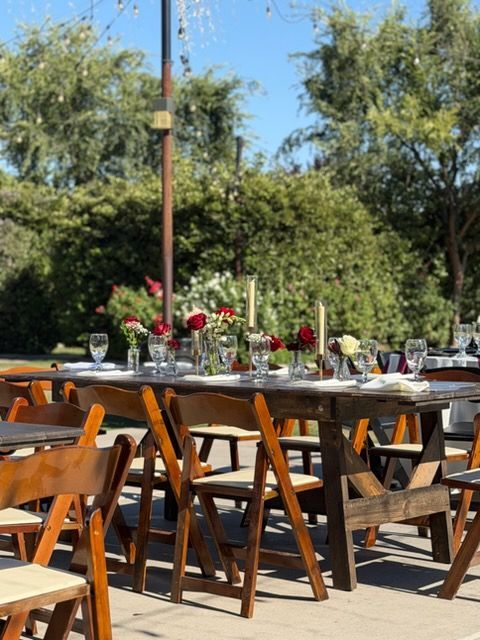 Outdoor dining table set with white tablecloth, wooden chairs, glasses, and red flowers in a garden.