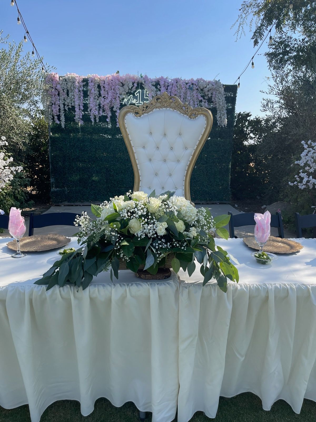 Elegant outdoor event table with white floral centerpiece and gold-trimmed throne chair on a decorated stage