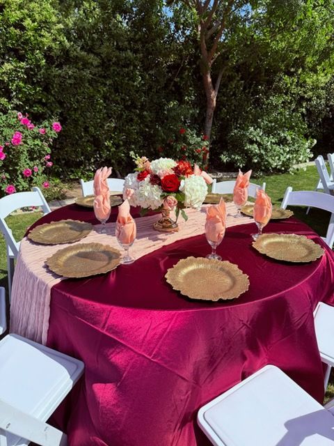 Outdoor round banquet table with burgundy tablecloth, gold chargers, floral centerpiece, and white chairs