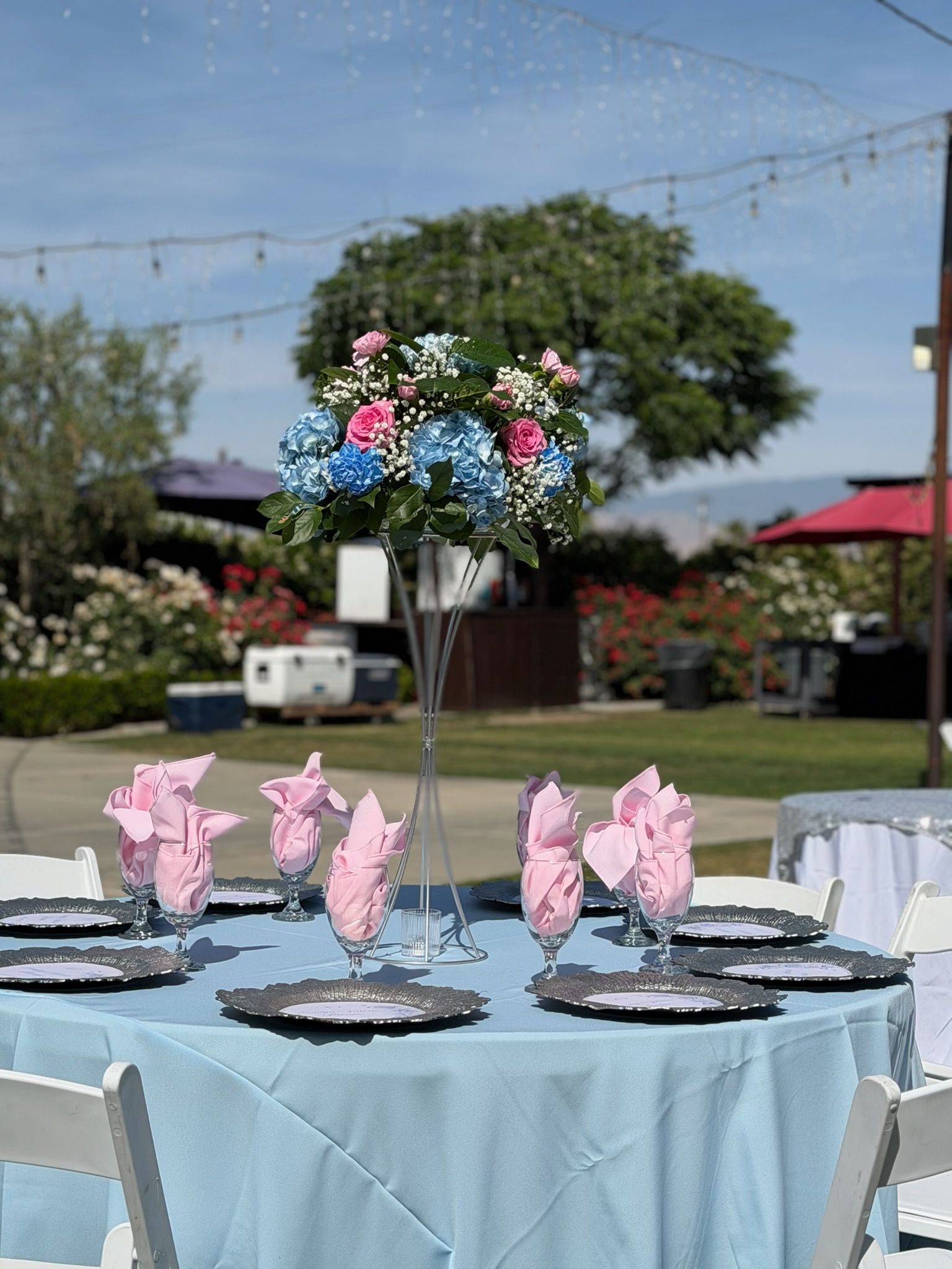 Outdoors banquet table with pink napkin decorations and a floral centerpiece under string lights.