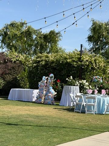 Outdoor garden reception setup with white-clothed tables, chairs, string lights, and floral decorations on a lawn