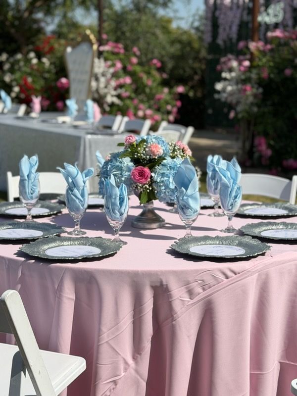 Pink-draped outdoor table set with blue napkins, floral centerpiece, and black chargers for a formal event
