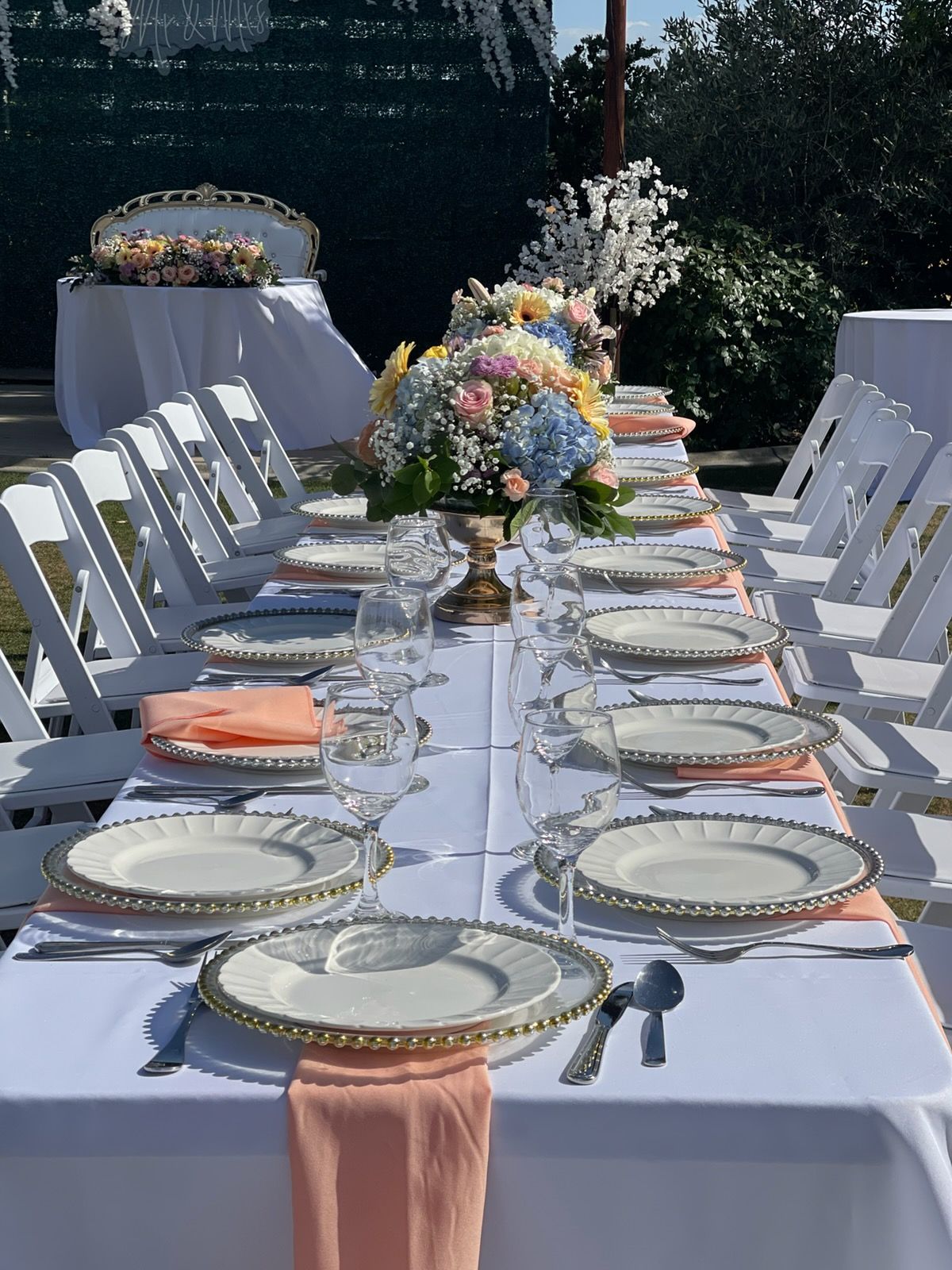 Elegant outdoor banquet table with white linens, gold-rimmed plates, peach napkins, and floral centerpieces