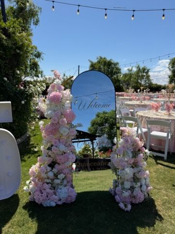 Pastel floral wedding welcome display with a sign, flowers, and pink chairs outdoors