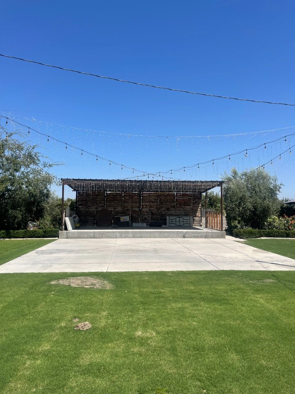 Outdoor stage with a concrete apron, grassy foreground, string lights, and trees under a clear blue sky