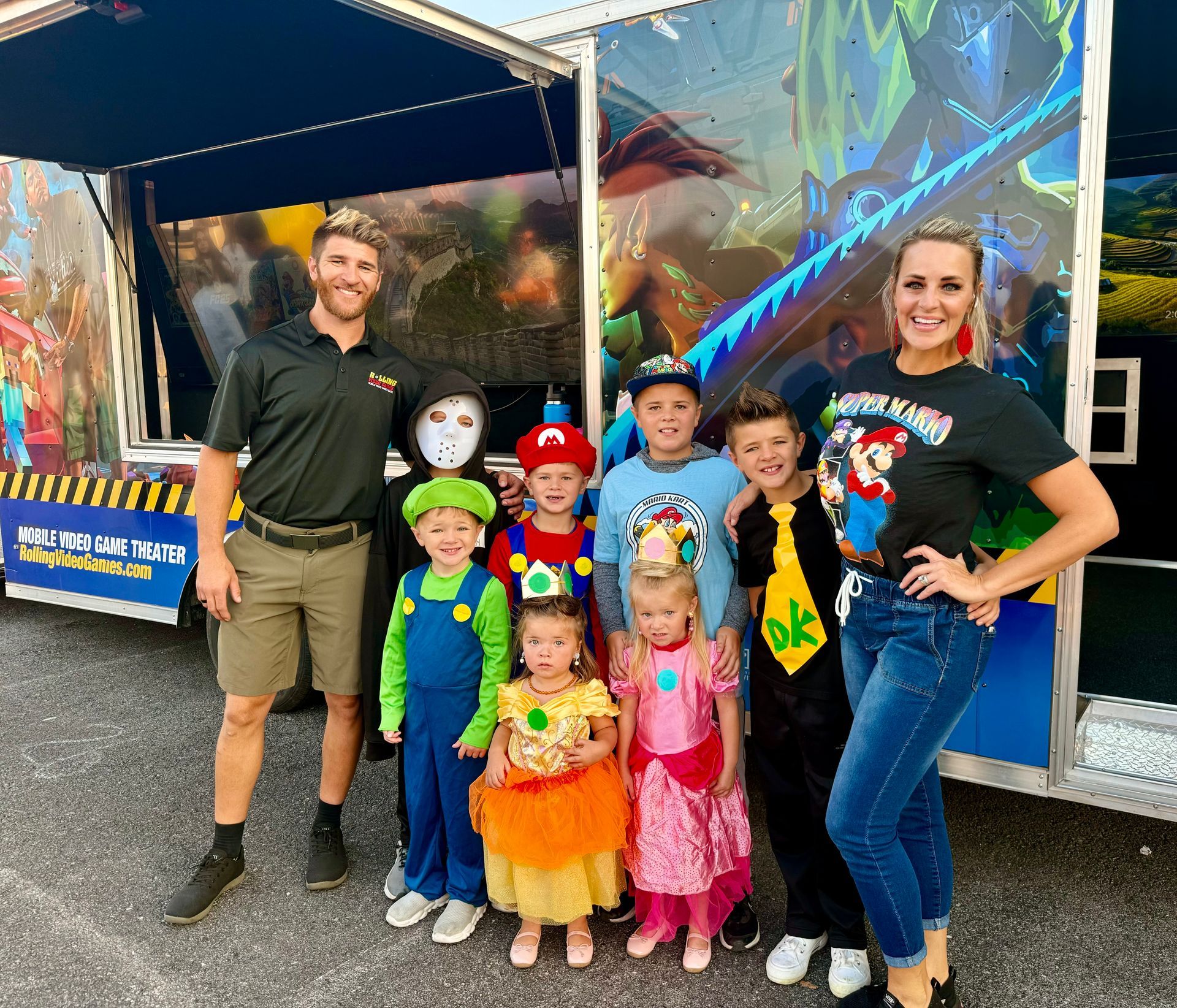 A group of adults and children dressed in colorful Super Mario-themed costumes pose together in front of a game trailer.