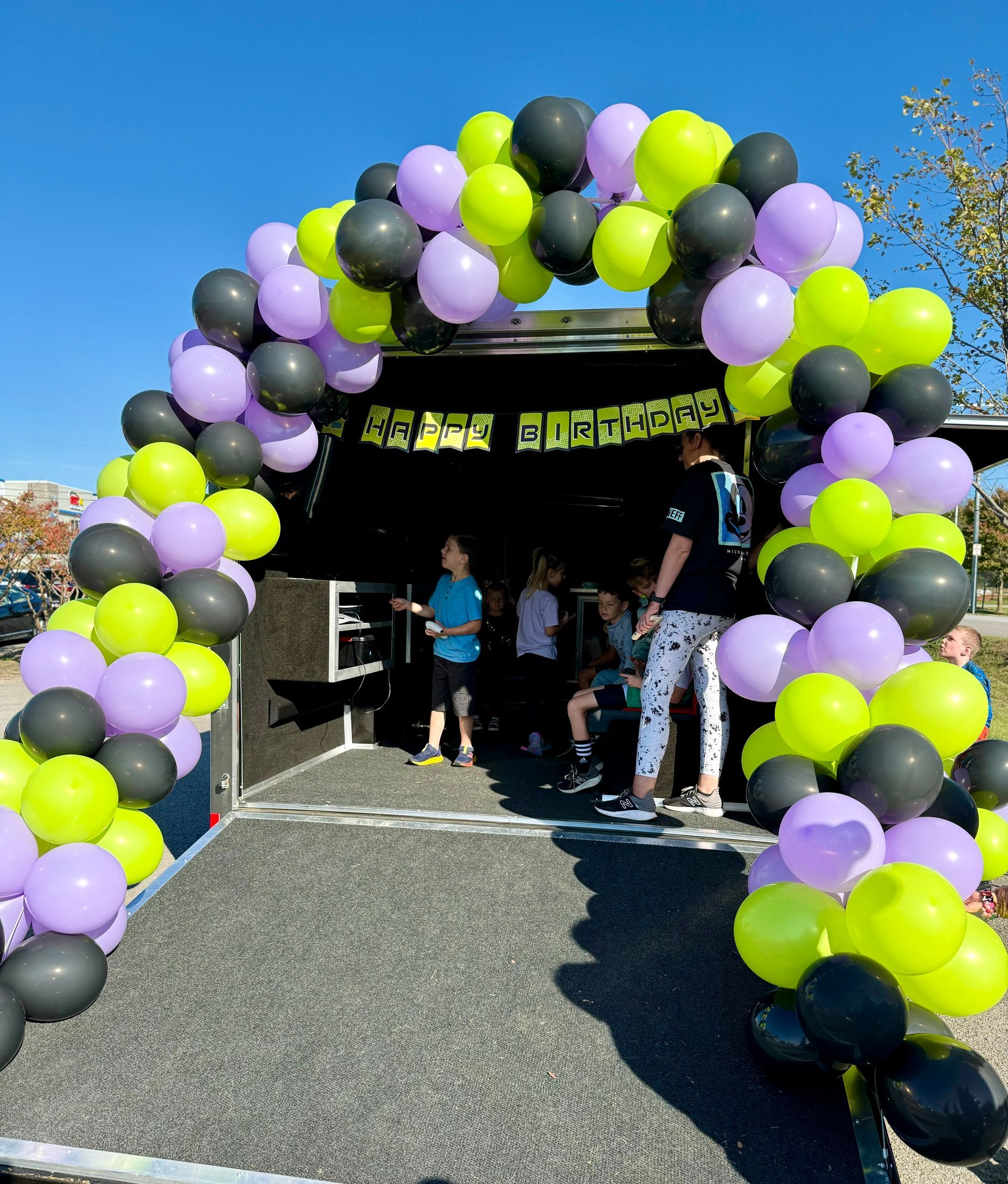 A trailer entrance decorated with an arch of black, neon green, and light purple balloons, with people inside.