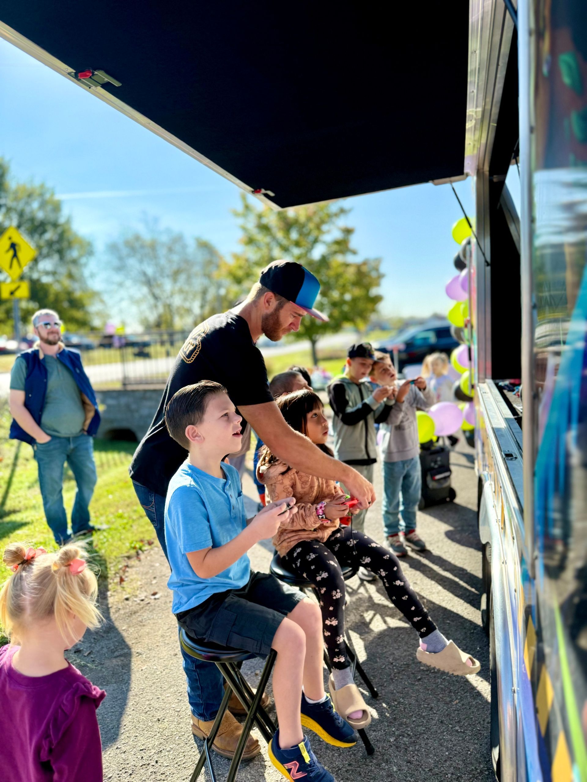 A person leans from a service truck to hand treats to children seated on stools outside on a sunny day.