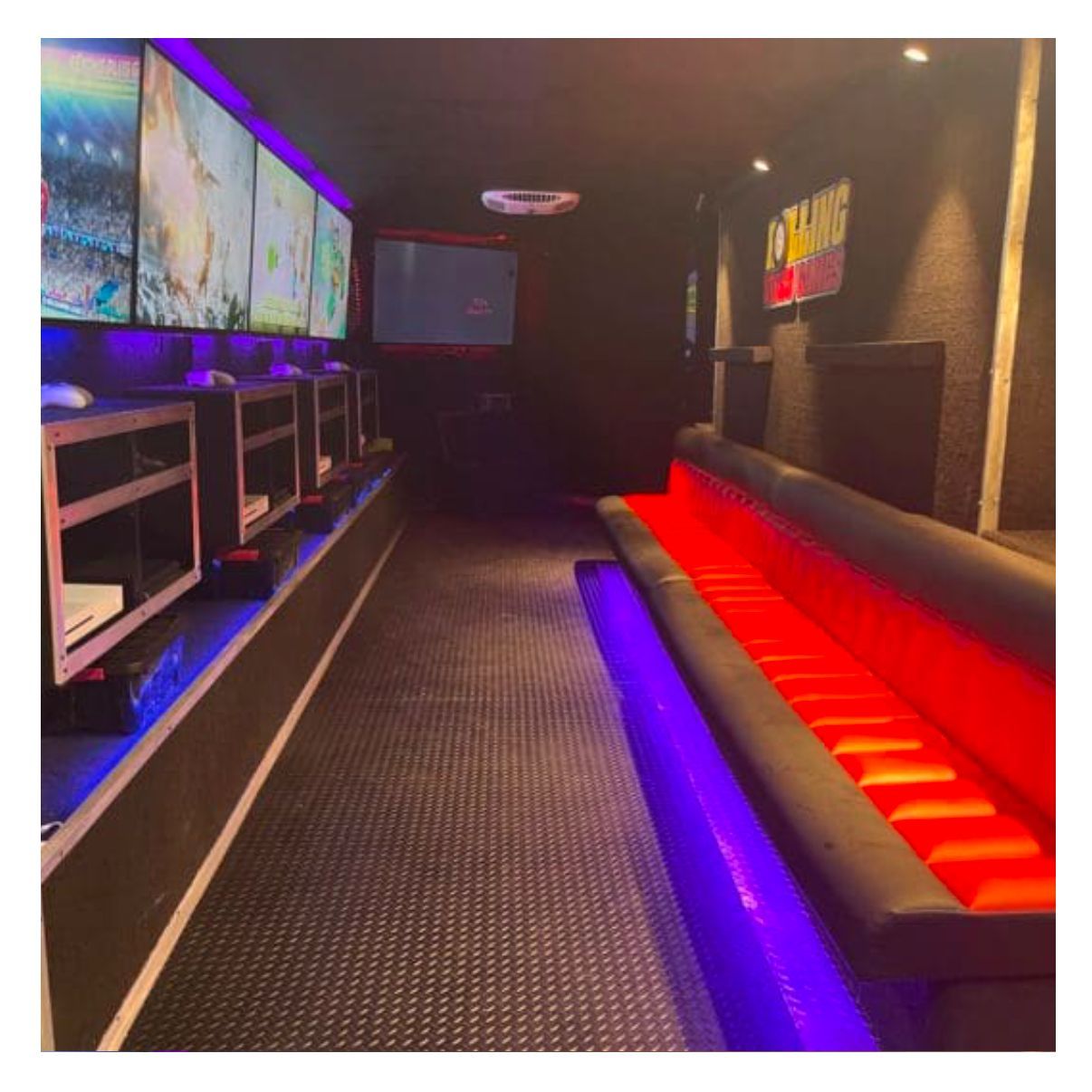Interior of a mobile gaming trailer with multiple wall-mounted screens, console stations, and long bench seating.
