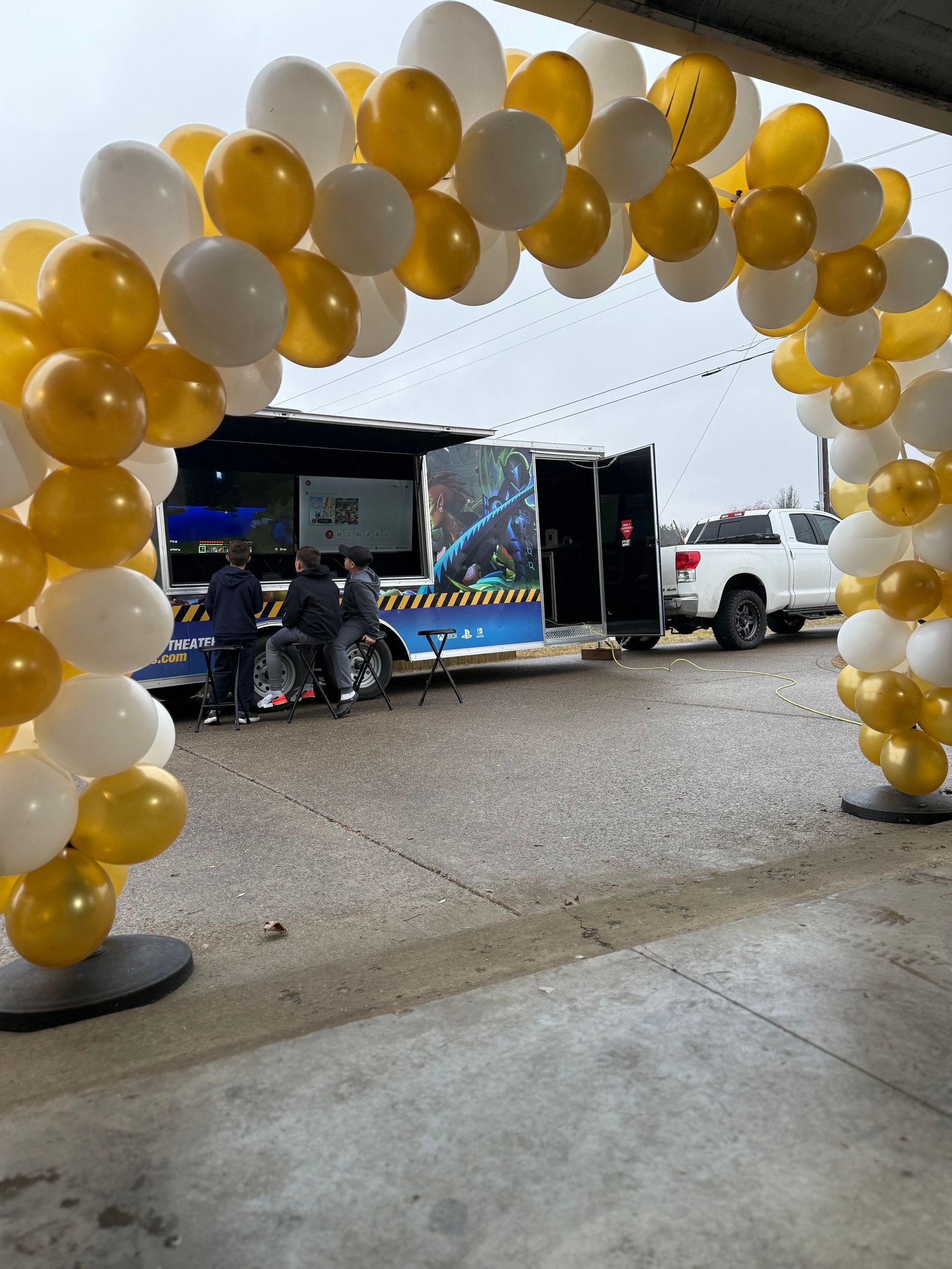 A balloon arch in white and gold stands outdoors, framing a view of a mobile event trailer and a parked white truck.